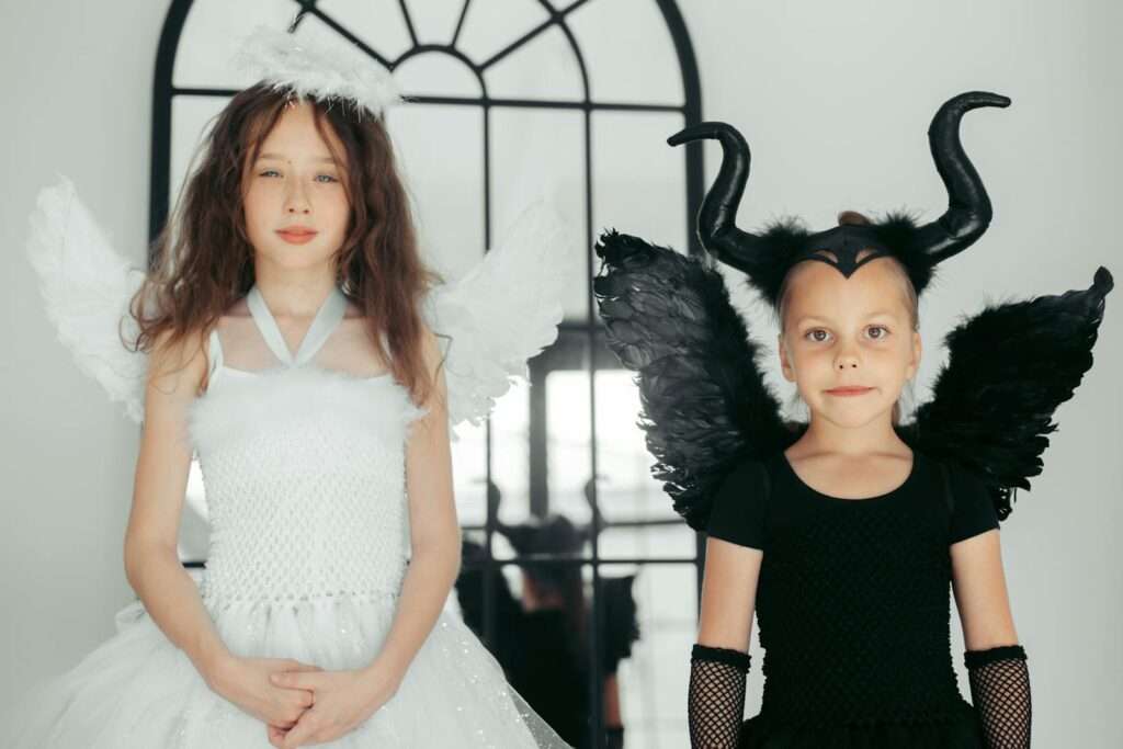 Two girls dressed as angel and devil pose indoors, a charming and playful exploration of duality.